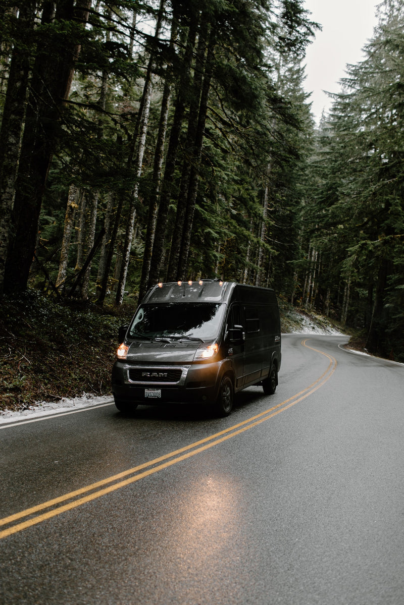 Dark RV rental van with lights on, navigating a wet, winding forest road surrounded by tall evergreens. Ideal for adventure. — photo 19