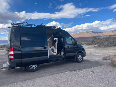 Black rv rental camper van with open side door and hammock, parked in a desert scene with mountains and clouds.