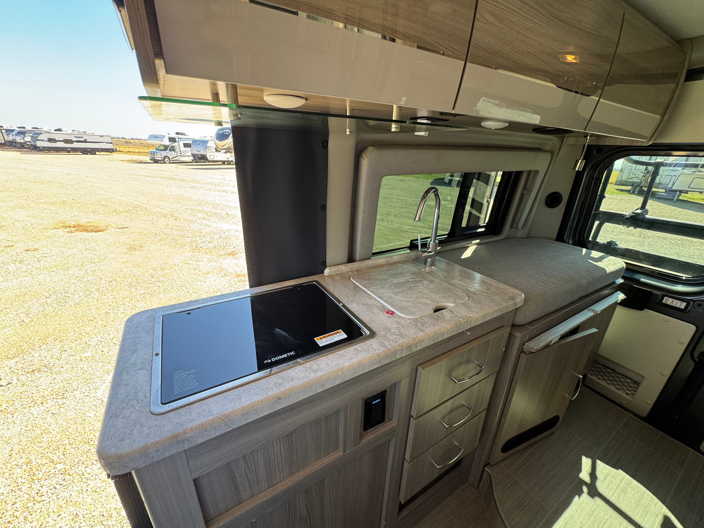 View of an RV rental's compact kitchen area, featuring a Dometic cooktop, sink, cabinets, and drawers. Other RVs are seen outside. — photo 9