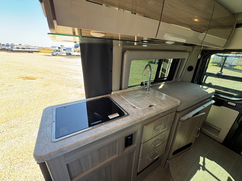 View of an RV rental's compact kitchen area, featuring a Dometic cooktop, sink, cabinets, and drawers. Other RVs are seen outside. — photo 9