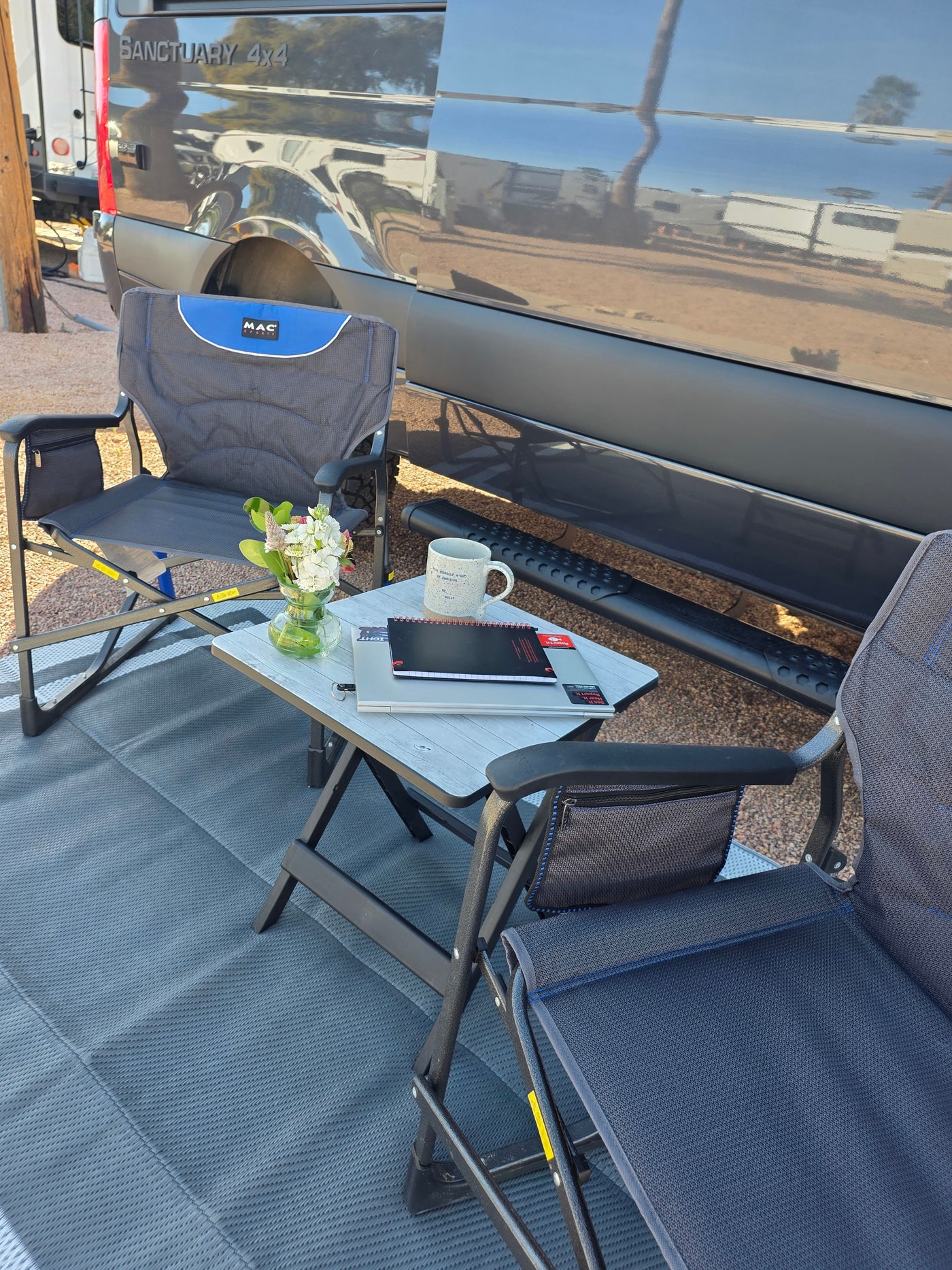 Relaxing outside a "Sanctuary 4x4" RV rental. Two camping chairs and a table with flowers, a mug, and notebooks, all on a gray mat. — photo 18