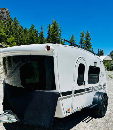 White Luna Rover camper rv rental parked on gravel. Features a black front cover, black wheels, against green trees & blue sky.