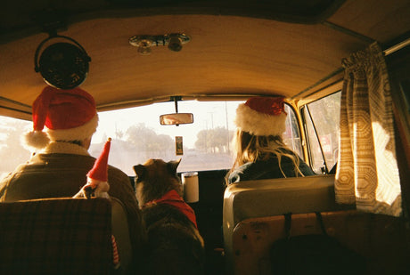 Inside a vintage RV rental: two people in Santa hats, a dog, and a festive road trip adventure. Warm light fills the cabin.