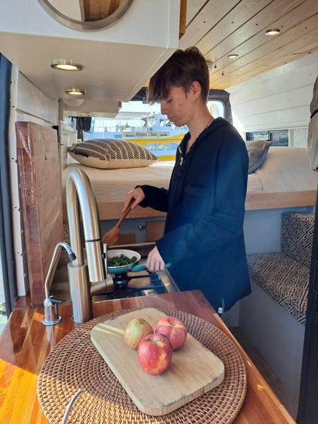 A person cooks in a modern RV rental, featuring a compact kitchen with wood accents and a cozy bed area.