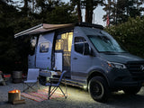 This grey Mercedes Sprinter camper van RV rental sets up a cozy night. Illuminated awning, open interior & fire pit glow brightly.