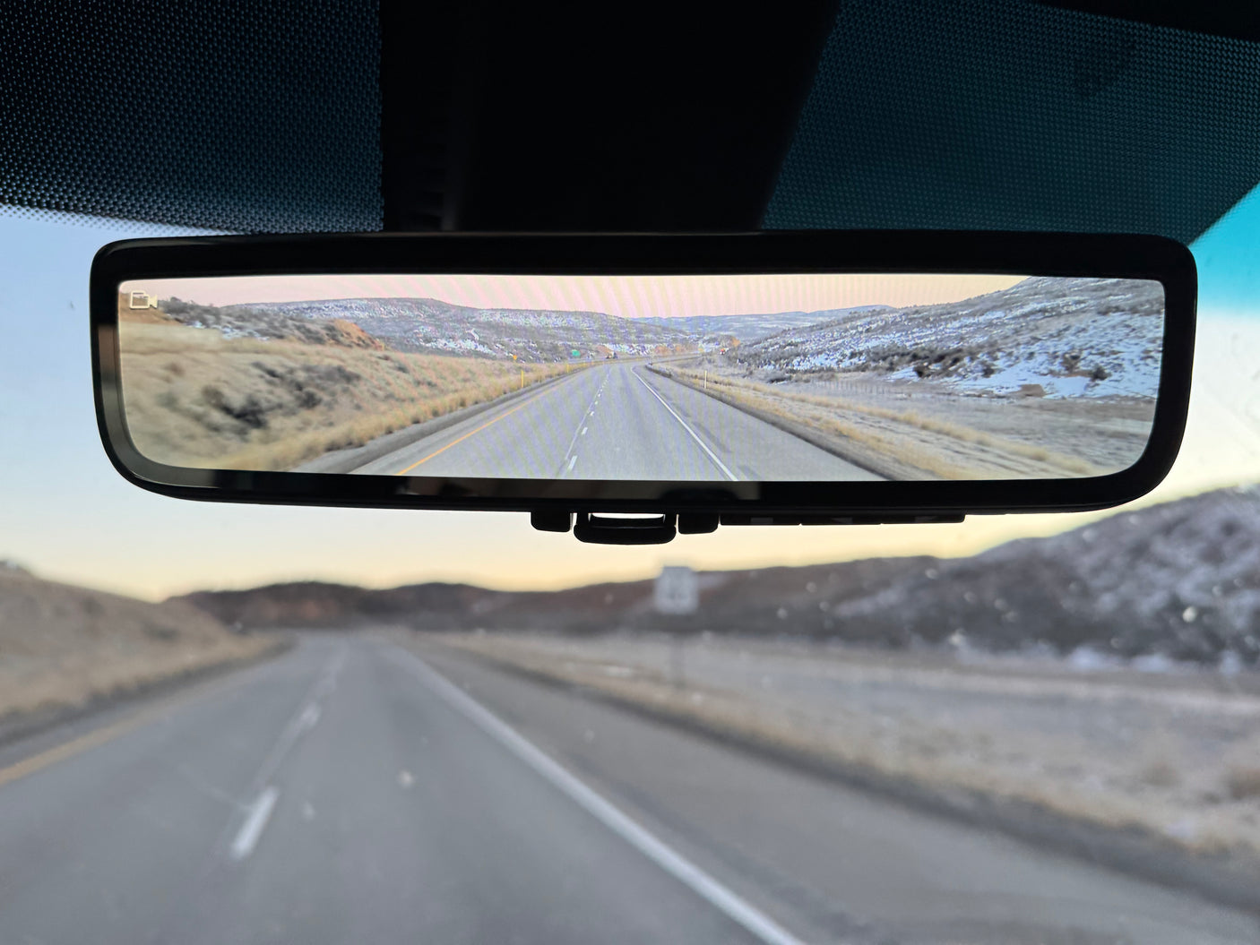 RV rental's digital rearview mirror displays a highway winding through snowy mountains. The real road is also visible through the windshield.