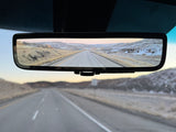 RV rental's digital rearview mirror displays a highway winding through snowy mountains. The real road is also visible through the windshield.