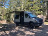 This is an RV rental, a grey Mercedes Sprinter camper van, parked with its awning extended in a sunny forest setting.
