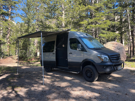 This is an RV rental, a grey Mercedes Sprinter camper van, parked with its awning extended in a sunny forest setting.