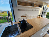 Stylish RV rental kitchen with sink, induction cooktop, and wood counter. Green landscape outside.