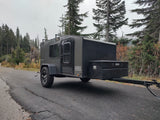 Compact Hiker Trailer RV rental parked on a mountain road, ready for adventure amidst tall evergreen trees.
