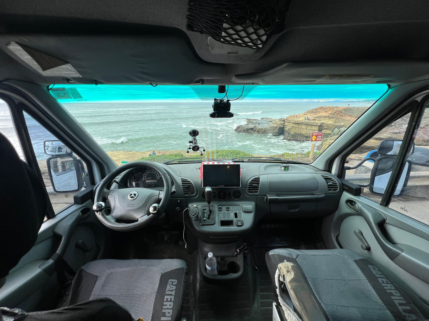 RV rental interior: driver's seat view of a stunning ocean vista from a Mercedes van. Coastal cliffs and dashboard visible.