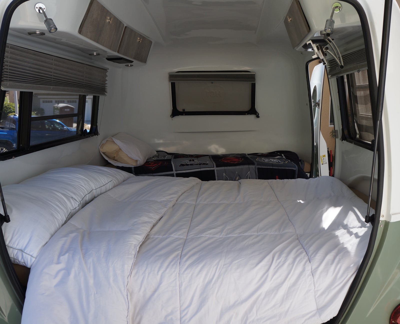 Compact rv rental bedroom with a comfy bed, white duvet, pillows, overhead cabinets, and windows with blinds. Ideal for travel.