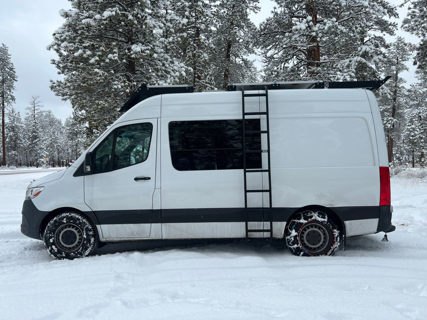 White **RV rental** van in a snowy winter forest, with tire chains on the rear wheel for traction.
