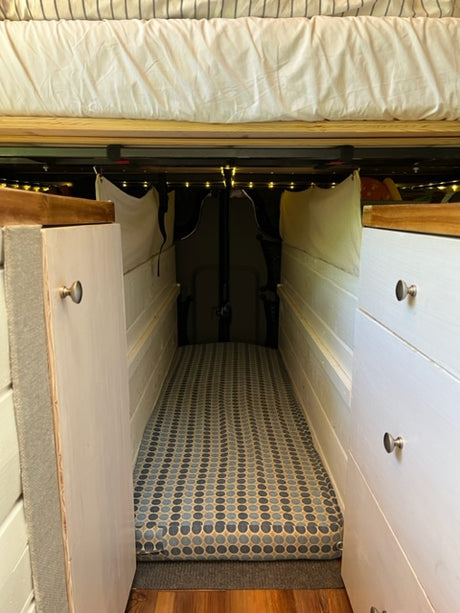 This RV rental interior features a cozy sleeping area, cabinet storage, patterned cushion, and decorative string lights.