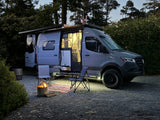 An RV rental parked at dusk, illuminated inside & out, with an open side, awning, fire pit, chairs, and rug for a cozy camp.