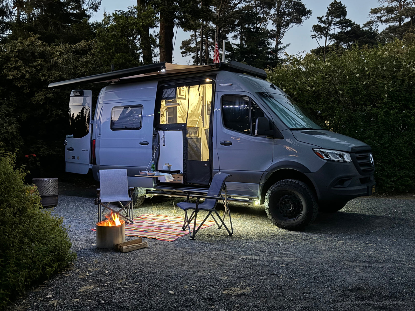 An RV rental parked at dusk, illuminated inside & out, with an open side, awning, fire pit, chairs, and rug for a cozy camp.