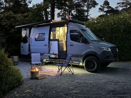 An RV rental parked at dusk, illuminated inside & out, with an open side, awning, fire pit, chairs, and rug for a cozy camp.