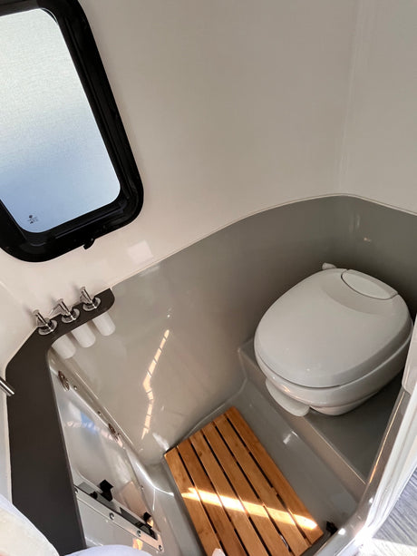 Compact RV rental wet bath featuring a toilet, shower area with a wooden mat, faucets, and a small window.