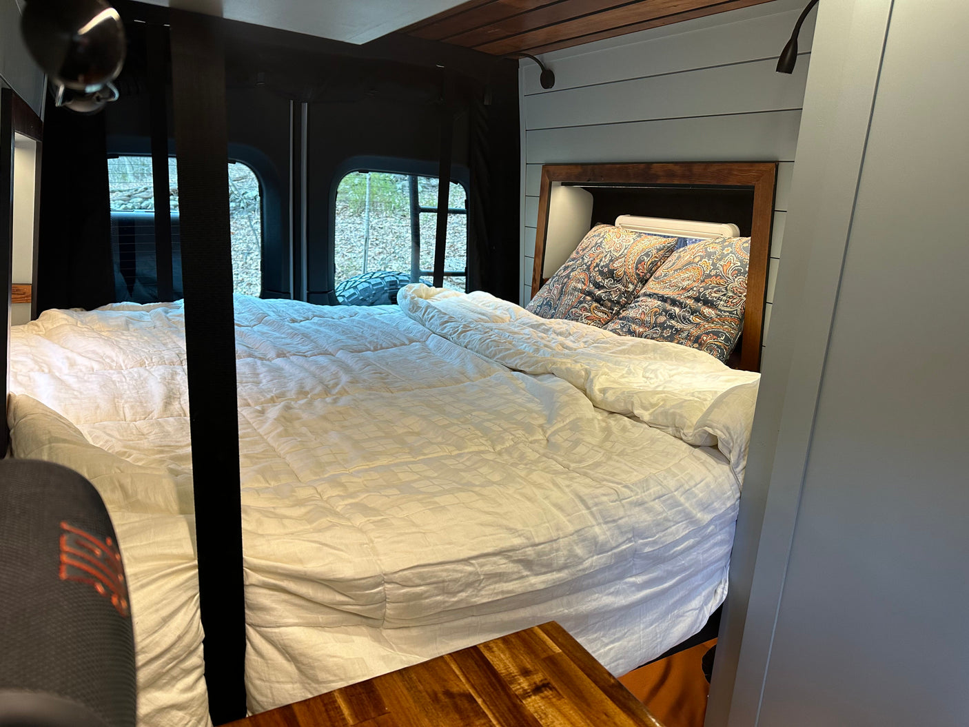 Cozy rv rental interior: bed with white bedding, patterned pillows, wooden headboard, light blue walls, and outside window views.