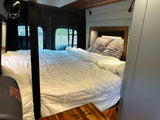 Cozy rv rental interior: bed with white bedding, patterned pillows, wooden headboard, light blue walls, and outside window views.