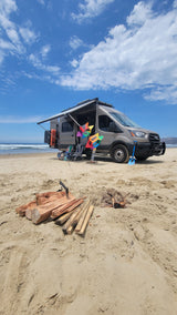 Colorful beach RV rental setup: a camper van with awning, pinwheels, campfire, and ocean.