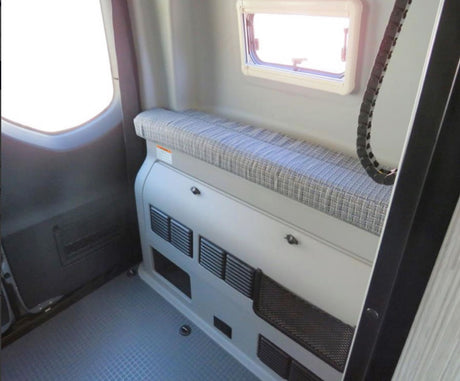 RV rental interior: long gray bench seat, upper window, and lower storage unit with vents on a textured floor.