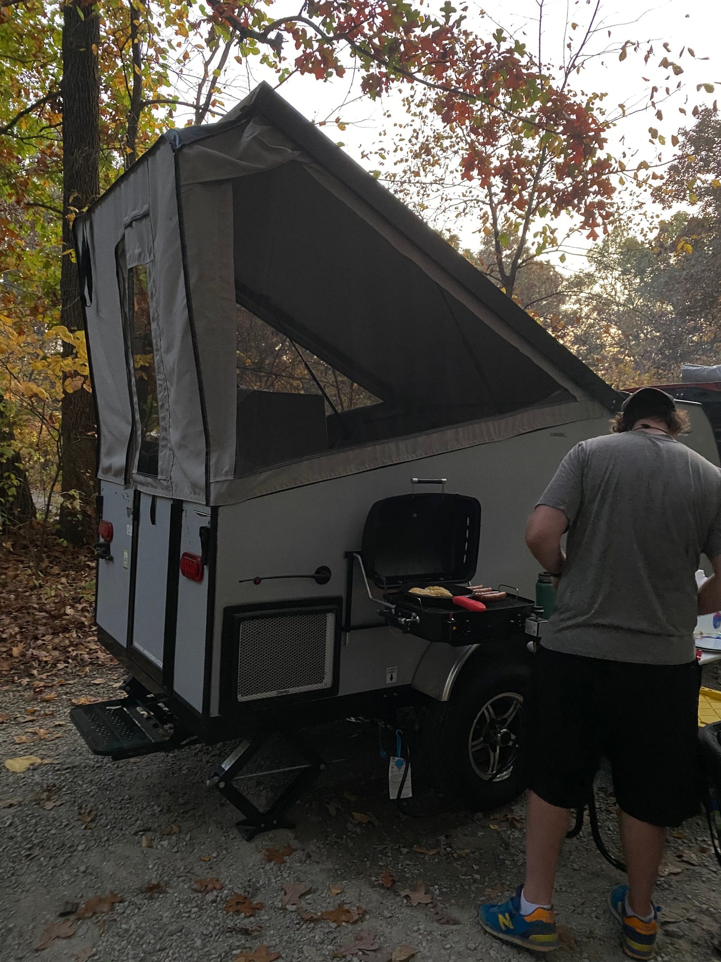 Autumn outdoor cooking scene: A person grills next to a pop-up RV rental, surrounded by fall foliage at a campsite.
