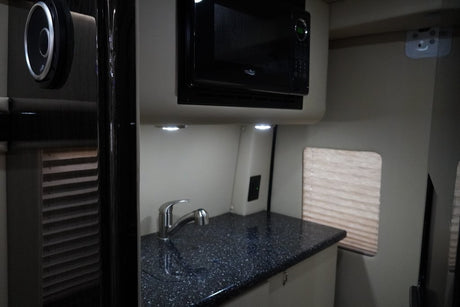 RV rental kitchen: microwave, sink, speckled countertop, and window with blinds. Brightly lit compact galley.