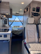 This RV rental features a cozy interior with kitchenette, seating, and a clear view of a tranquil lake from the open rear doors.