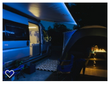 This lit campervan RV rental provides a cozy night camping setup. An extended awning illuminates the campsite with a tent and picnic table.