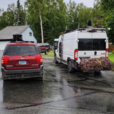 Explore with this **RV rental**! A white customized van & red SUV, loaded with travel gear, parked on a wet street.