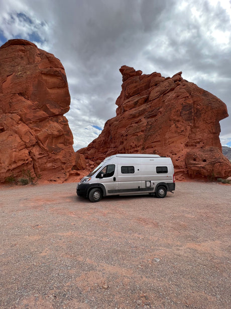 Silver camper van RV rental parked on a gravel path between towering red rock formations, under a cloudy desert sky.