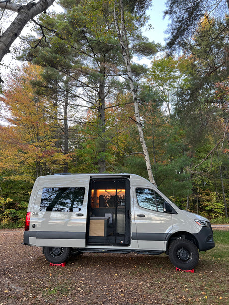 A light grey rv rental camper van, door open revealing a warm interior, parked among vibrant fall trees.