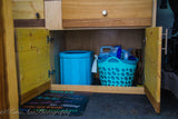 RV rental interior: Under a wooden cabinet, a blue collapsible bin, turquoise basket of supplies, and a 'rules' mat offer organized storage.