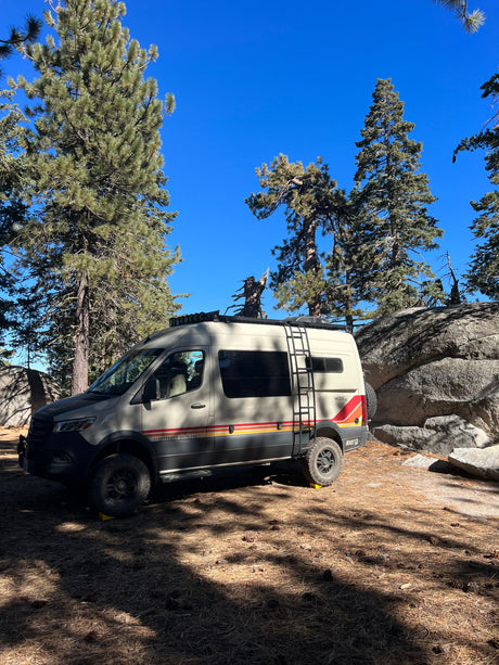 An adventure-ready camper van is parked among pine trees and large boulders, perfect for your next RV rental trip.