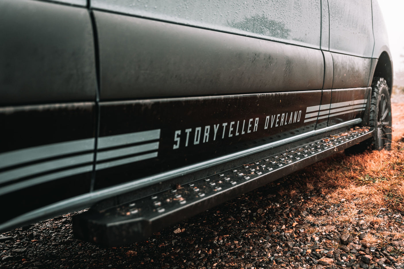 Side view of a dark Storyteller Overland camper van with rugged tires and a step, ideal for your next rv rental.