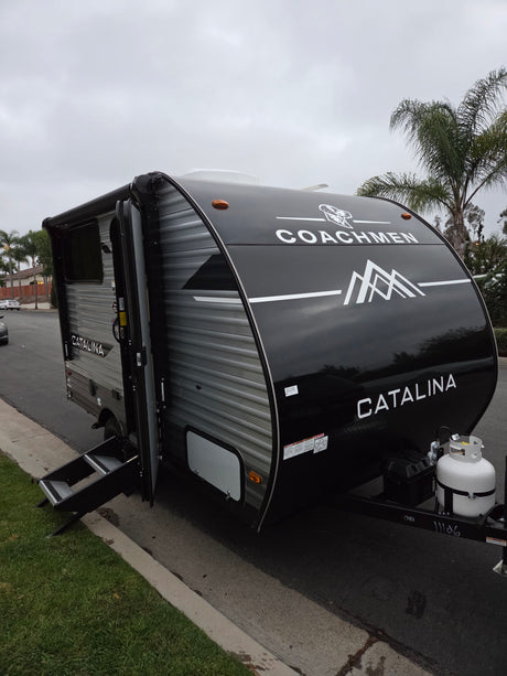 Black Coachmen Catalina travel trailer RV rental parked roadside with steps. Overcast day, palm trees in background.