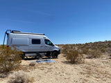Enjoying a desert RV rental adventure. Silver camper van with chairs & mat parked on sandy ground under clear blue sky.