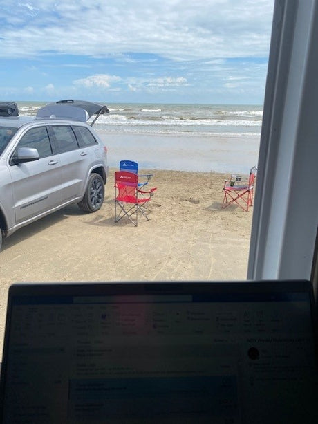RV rental view: laptop in foreground, silver SUV & chairs on beach, ocean waves beyond under cloudy sky.