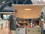 Man smiles, cooking at an integrated outdoor kitchen of an RV rental trailer, with a Yeti cooler. Perfect for camping adventures.