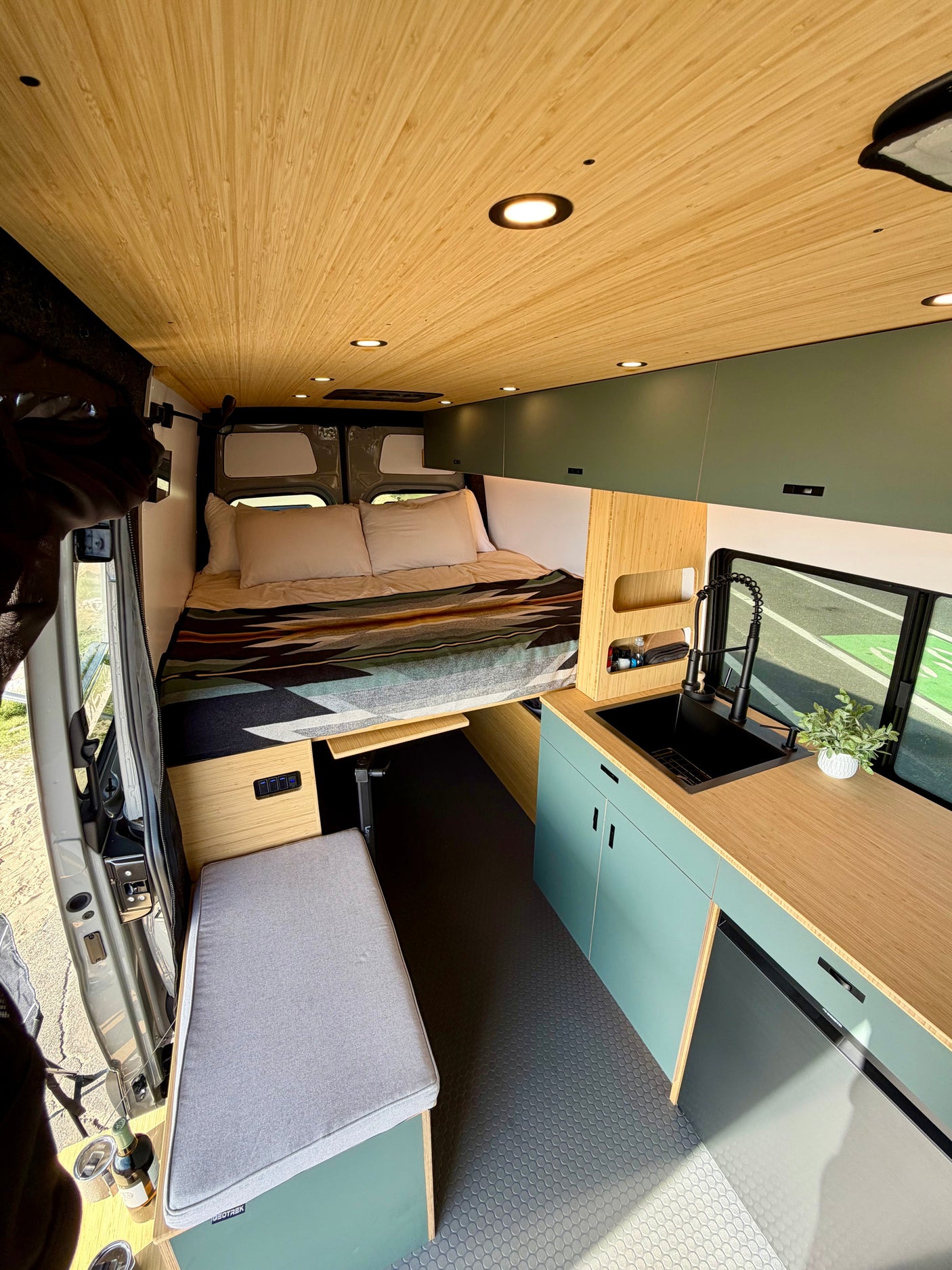 Modern rv rental camper van interior: wood ceiling, cozy bed, sleek kitchen with black sink, light green cabinets, and seating.