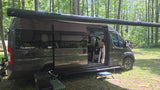 Enjoying the forest in a dark gray Winnebago Travato RV rental with its awning extended and door open, perfect for camping.