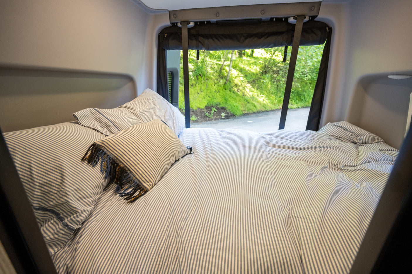 Cozy RV rental bed with striped bedding and a view of green nature outside, offering a comfortable night's sleep on the road.