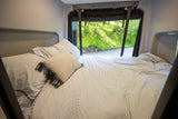 Cozy RV rental bed with striped bedding and a view of green nature outside, offering a comfortable night's sleep on the road.