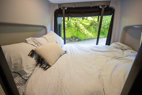 Cozy RV rental bed with striped bedding and a view of green nature outside, offering a comfortable night's sleep on the road.
