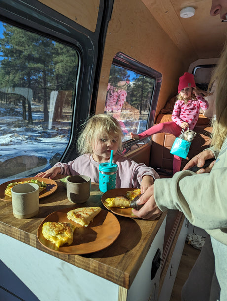 Family enjoys breakfast inside a cozy RV rental. Kids eat eggs & avocado with a snowy forest view.