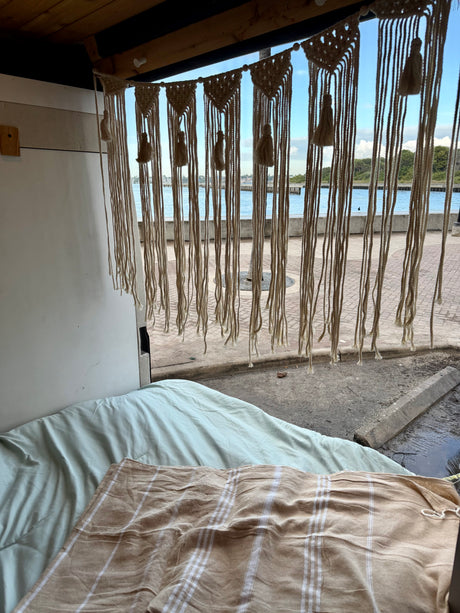 Cozy RV rental interior with a bed, macrame decor, and a scenic water view from the open back.
