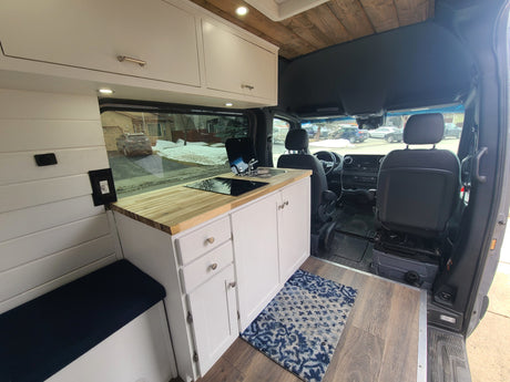 Bright custom RV rental interior: white cabinets, wood counter, cooktop, sink, comfy bench. A modern camper van conversion.