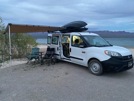 This campervan RV rental is parked lakeside with an awning & chairs, perfect for a cozy camping adventure. Stunning views!
